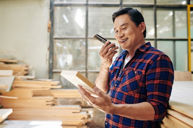 Sorridente carpentiere asiatico che guarda un mattone di legno in mano durante la registrazione di un messaggio vocale al cliente