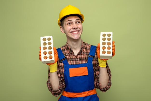 Sorridente cercando lato giovane costruttore maschio che indossa l'uniforme con i guanti che tengono i mattoni