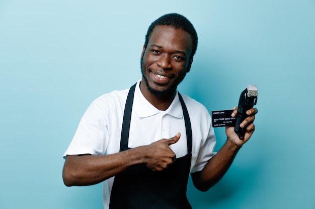 Sorridente che mostra i pollici in su tenendo la carta con le tosatrici giovane barbiere afroamericano in uniforme isolato su sfondo blu