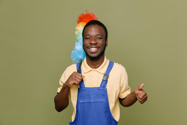 Sorridente che mostra i pollici in su tenendo pipidaster giovane maschio pulitore afroamericano in uniforme isolato su sfondo verde