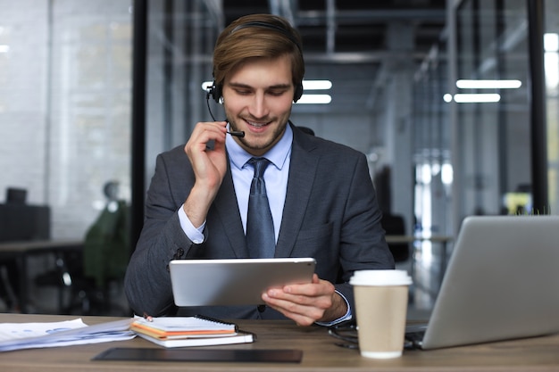 Sorridente consulente aziendale maschio con le cuffie seduto in un ufficio moderno, videochiamata guardando lo schermo del laptop. Assistenza clienti dell'agente del servizio clienti dell'uomo che parla di chat online.