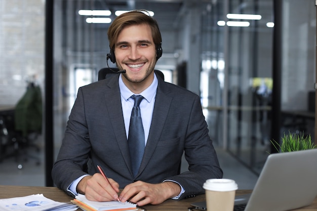 Sorridente consulente aziendale maschio con le cuffie seduto in un ufficio moderno, videochiamata guardando lo schermo del laptop. Assistenza clienti dell'agente del servizio clienti dell'uomo che parla di chat online.