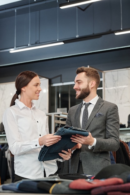 Sorridente consulente del negozio di abbigliamento che dà i jeans all'uomo mentre lo assiste nella scelta dei vestiti in negozio