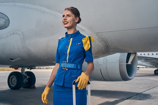 Sorridente contenta giovane assistente di volo femminile in guanti di pelle appoggiata alla maniglia retrattile del bagaglio