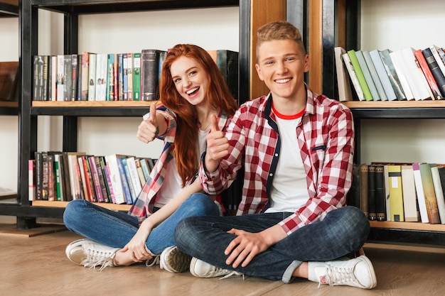 Sorridente coppia adolescente seduto su un pavimento presso lo scaffale per libri