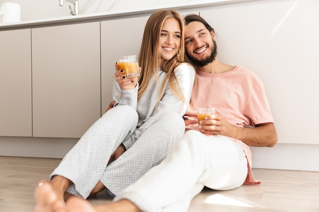 Sorridente coppia adorabile che si abbraccia mentre è seduta sul pavimento della cucina, bevendo succo d'arancia