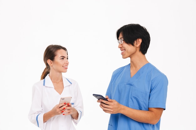 Sorridente coppia di medici indossando uniforme in piedi isolato sopra il muro bianco, utilizzando i telefoni cellulari