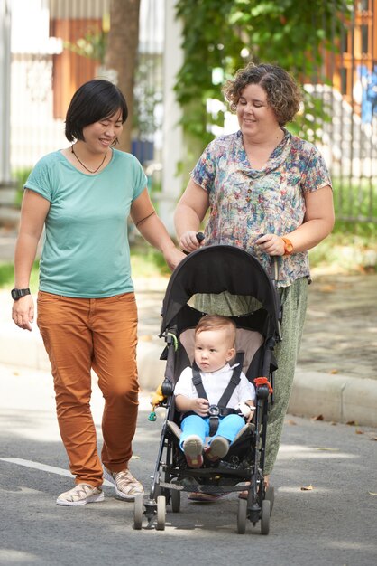 Sorridente coppia lesbica multietnica che cammina all'aperto con il piccolo figlio in carrozzina