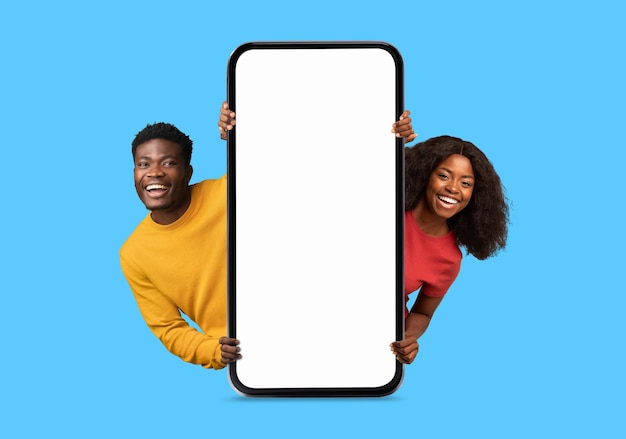 Sorridente coppia millenaria afroamericana che fa capolino da un enorme smartphone con schermo vuoto consiglia il blog