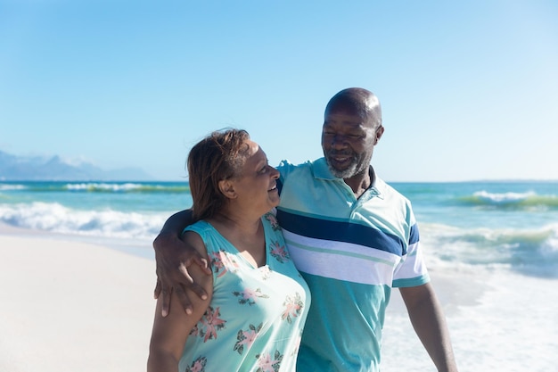 Sorridente coppia senior afroamericana che cammina con il braccio intorno alla spiaggia in una giornata di sole