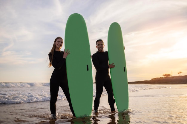 Sorridente coppia sportiva in mute in posa con tavole da surf sulla spiaggia al tramonto