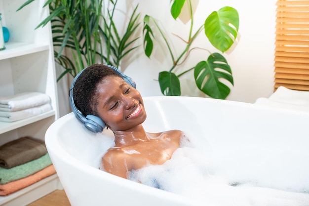 Sorridente donna africana attraente che indossa le cuffie rilassandosi e ballando nel bagno di schiuma in bagno