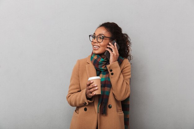 Sorridente donna africana che indossa cappotto in piedi, parlando al telefono cellulare, tenendo la tazza di caffè