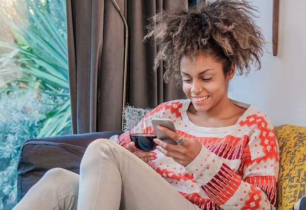 Sorridente donna afro-americana seduta sul divano e la lettura di messaggi di testo. Sorridente giovane donna a casa che si distende sul divano, sta utilizzando uno smartphone e il testo, la tecnologia e la comunicazione concetto