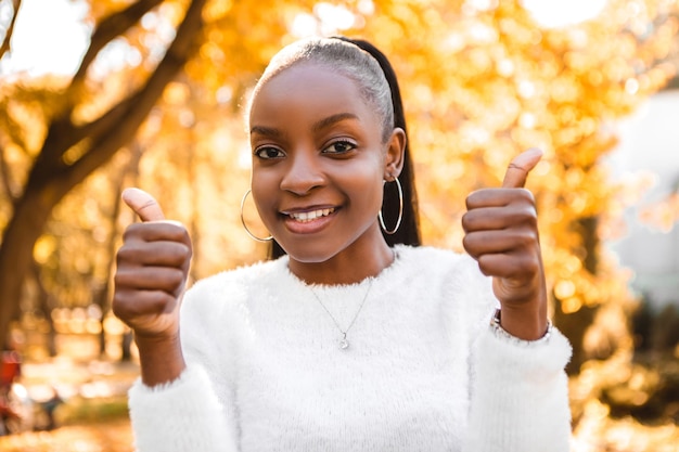 Sorridente donna afroamericana in piedi nel parco cittadino, divertendosi, mostrando il pollice in su come