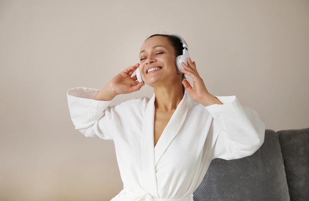 Sorridente donna afroamericana o di razza mista in accappatoio bianco con le cuffie che si divertono ad ascoltare la musica.