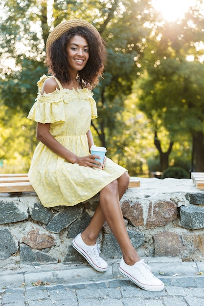 Sorridente donna americana 20s che indossa cappello di paglia e vestito, seduta su una panchina nel parco e bevendo caffè da asporto
