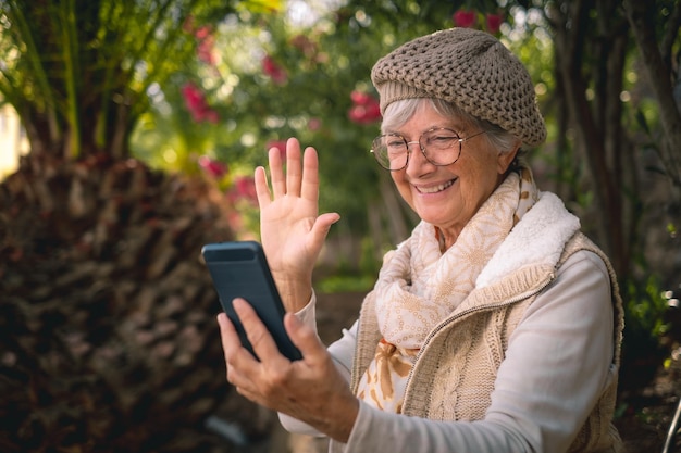 Sorridente donna anziana anziana seduta nel parco in chat video con il telefono cellulare salutando Bella signora in pensione all'aperto seduta all'ombra degli alberi