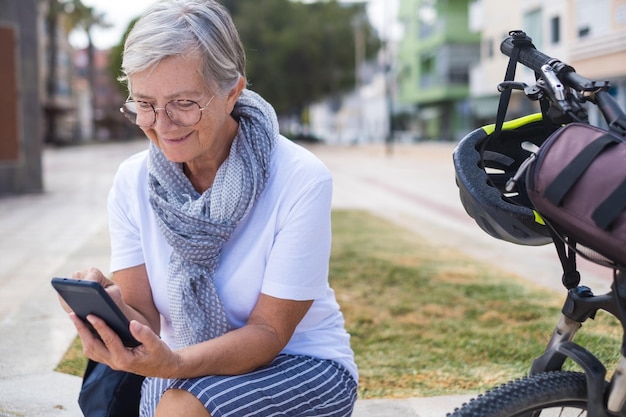 Sorridente donna anziana caucasica vestita casual seduta all'aperto vicino alla sua bicicletta utilizzando il telefono cellulare per la messaggistica