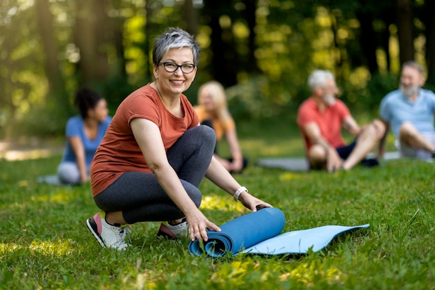 Sorridente donna anziana che rotola tappetino yoga dopo una lezione di gruppo all'aperto