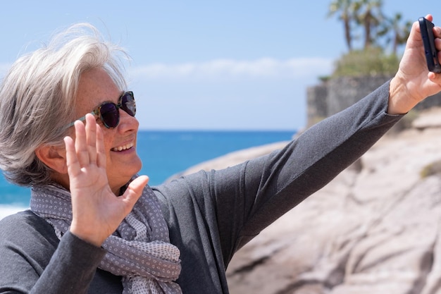 Sorridente donna anziana onda alla telecamera con videochiamata sul telefono cellulare all'aperto in vacanza in mare Felice donna anziana in piedi sulla spiaggia parlare utilizzando moderne tecnologie e connessione wireless