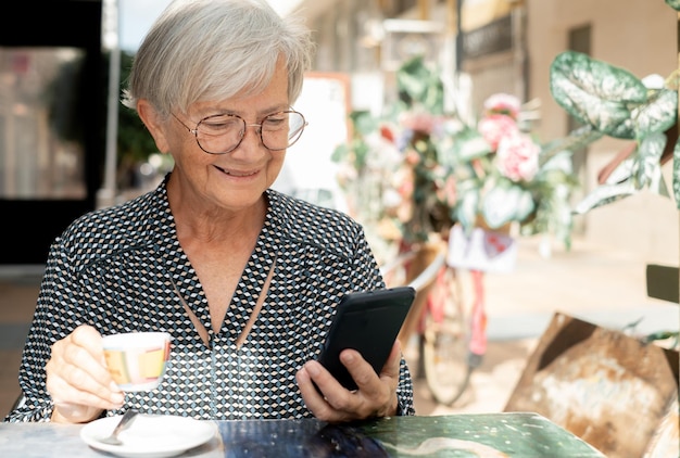 Sorridente donna anziana seduta al caffè all'aperto utilizzando il suo smartphone godendo di una pausa caffè
