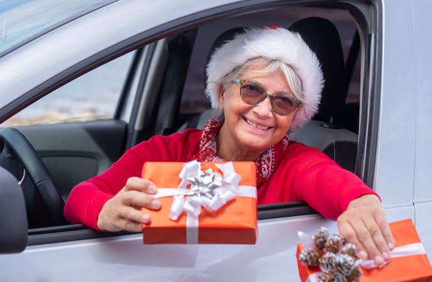 Sorridente donna anziana vestita di rosso si sporge dal finestrino della macchina indossando un cappello di Natale e tenendo in mano i regali di Natale Christmas