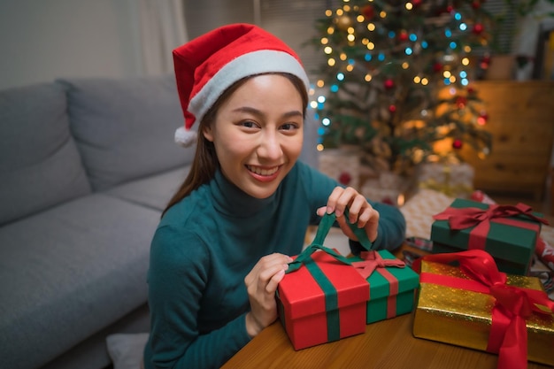 Sorridente donna asiatica che indossa cappello da Babbo Natale e confezione regalo sul tavolo per Natale guarda la telecamera con l'albero di Natale sullo sfondo