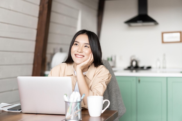 Sorridente donna asiatica che lavora al computer portatile da casa sognando qualcosa con la faccia felice che pensa mentre...