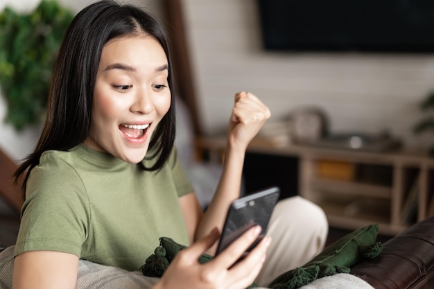 Sorridente donna asiatica che sembra soddisfatta mentre fa la pompa a pugno e legge il cellulare seduto a casa e...