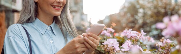 Sorridente donna asiatica dai capelli grigi usa il cellulare in piedi sulla terrazza all'aperto