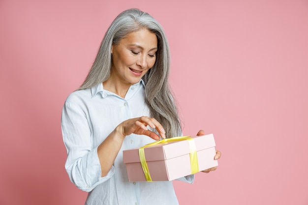 Sorridente donna asiatica di mezza età apre una confezione regalo con un nastro su sfondo rosa