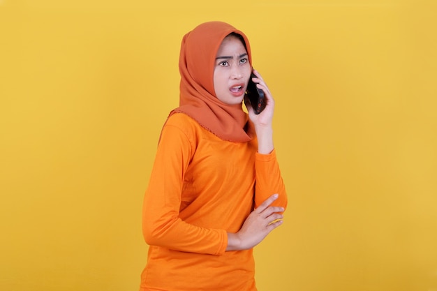 Sorridente donna asiatica felice con espressione sussurrata isolata su sfondo giallo chiaro banner che indossa l'hijab, parlando con il telefono cellulare