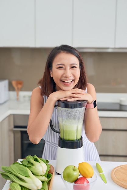 Sorridente donna asiatica vegana che prepara un frullato con frutta e verdura in cucina