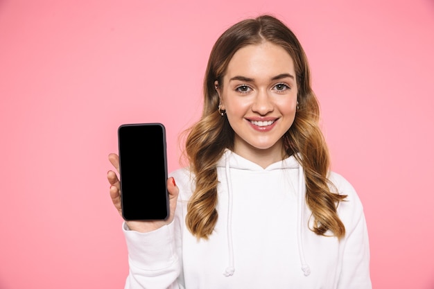 Sorridente donna bionda che indossa in abiti casual che mostra lo schermo dello smartphone vuoto e guardando la parte anteriore sul muro rosa