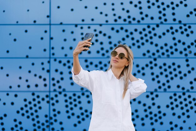 Sorridente donna bionda in occhiali da sole scatta una foto selfie contro un muro blu con puntini neri