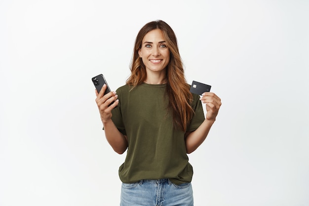 Sorridente donna bruna che mostra la sua carta sconto di credito, tenendo in mano il telefono cellulare dello smartphone, guardando soddisfatta davanti