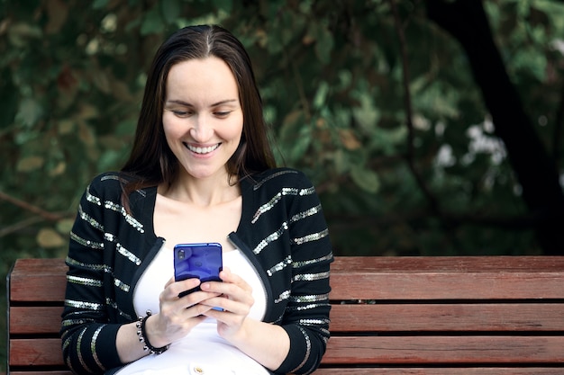 Sorridente donna bruna con lo smartphone nelle sue mani è seduto mandare SMS sulla panchina nel parco estivo, concetto di comunicazione