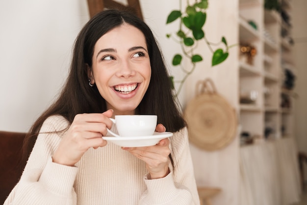 sorridente donna bruna tenendo la tazza e bere il caffè mentre si fa colazione in cucina a casa