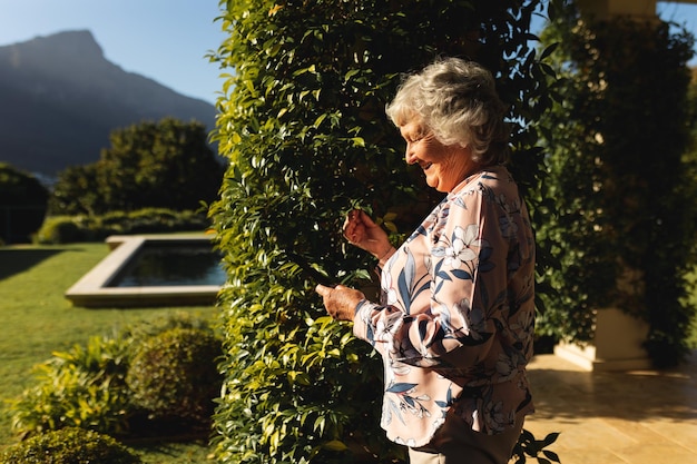 Sorridente donna caucasica anziana che usa lo smartphone nel giardino soleggiato. stile di vita pensionistico di lusso, trascorrere del tempo da solo a casa.