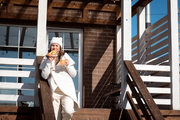 Sorridente donna caucasica felice in abiti bianchi che mangia pizza sul portico della casa suburbana