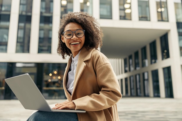 Sorridente donna d'affari africana che lavora al computer portatile mentre si siede fuori sullo sfondo dell'ufficio