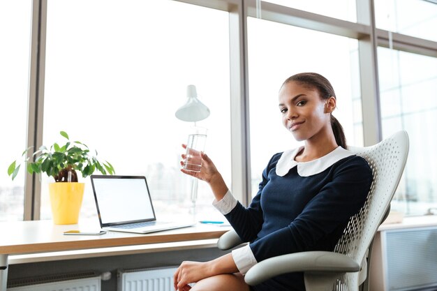 Sorridente donna d'affari afro in abito seduta al tavolo vicino alla finestra con una tazza d'acqua in mano e guardando la telecamera in ufficio