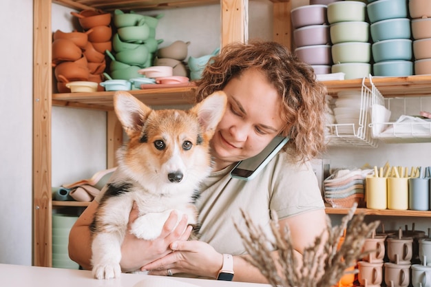 Sorridente donna d'affari concentrata che parla con uno smartphone che tiene un simpatico cane corgi in officina Negozio di piatti per bambini in silicone