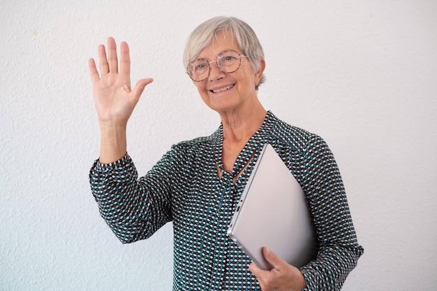 Sorridente donna d'affari matura in camicia e occhiali isolati su sfondo bianco tenendo il laptop sotto il braccio mentre agitando la mano