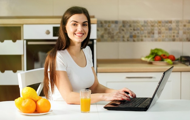 Sorridente donna dai capelli scuri in chat sul laptop con succo durante la chat positiva atmosfera rilassante a casa dolce