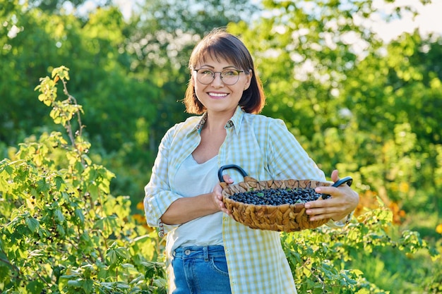 Sorridente donna di mezza età che tiene un cesto di ribes nero maturo Raccolta del ribes in giardino Coltivazione di bacche biologiche sane stagione estiva agricoltura concetto di cibo vitaminico