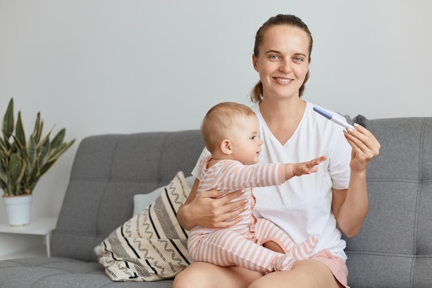 Sorridente donna felice che indossa una maglietta bianca in stile casual in posa con la figlia neonata e il test di gravidanza nelle mani, guardando la telecamera con un'espressione facciale positiva.