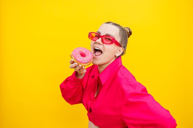 Sorridente donna incinta in camicia rosa con deliziose ciambelle dolci su sfondo giallo isolato Dolce cibo calorico durante la gravidanza Una donna incinta mangia pasticcini grassi