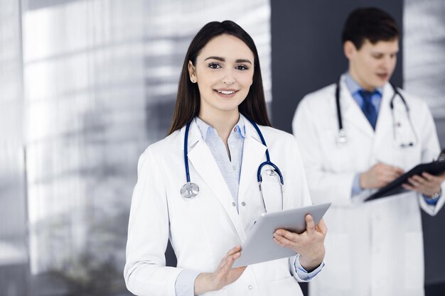 Sorridente donna-medico intelligente tiene in mano un tablet computer, mentre è in piedi in una clinica con un collega sullo sfondo. Ritratto di medico al lavoro. Servizio medico perfetto